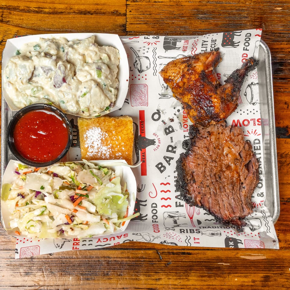 Barbecue platter with grilled chicken and ribs, potato salad, coleslaw, barbecue sauce, and cornbread on a wooden table.