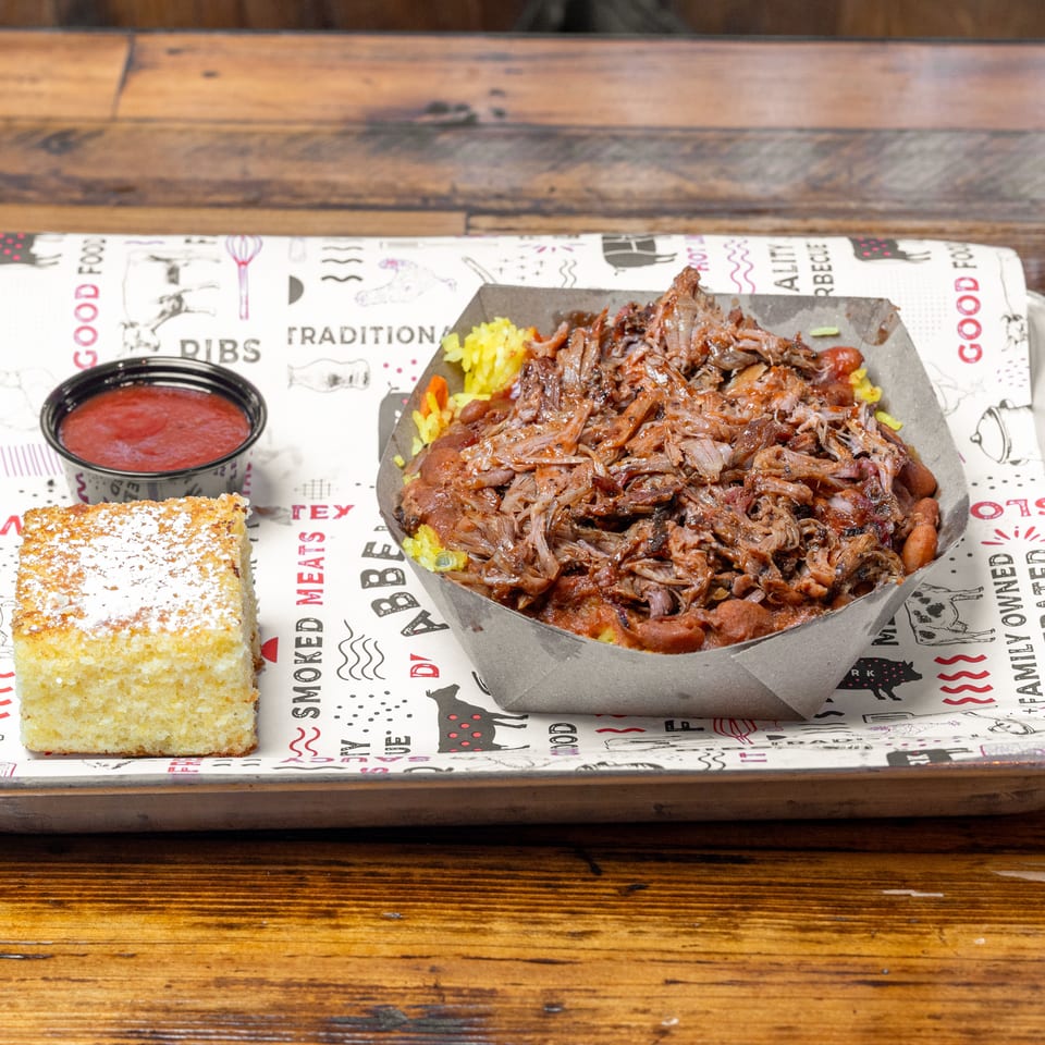 Tray of shredded barbecue meat over yellow rice with red sauce and powdered cornbread on a rustic wooden table.