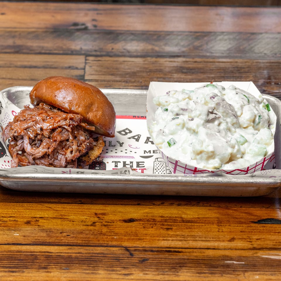 Pulled pork sandwich on a brioche bun with creamy potato salad, served on a metal tray atop a wooden table.