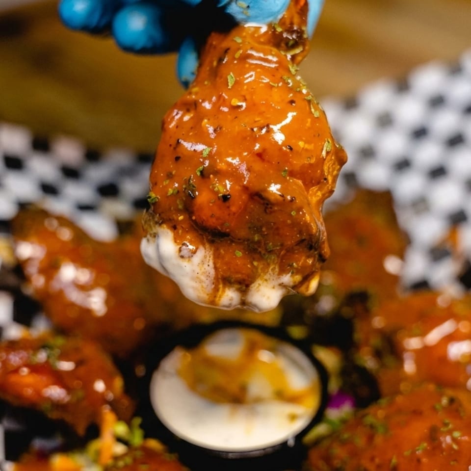Glazed chicken wing held above a basket of sauced wings with dipping sauce on checkered paper.