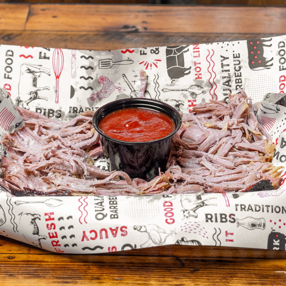Shredded barbecue pork served in a lined tray with a cup of red sauce on a wooden table.