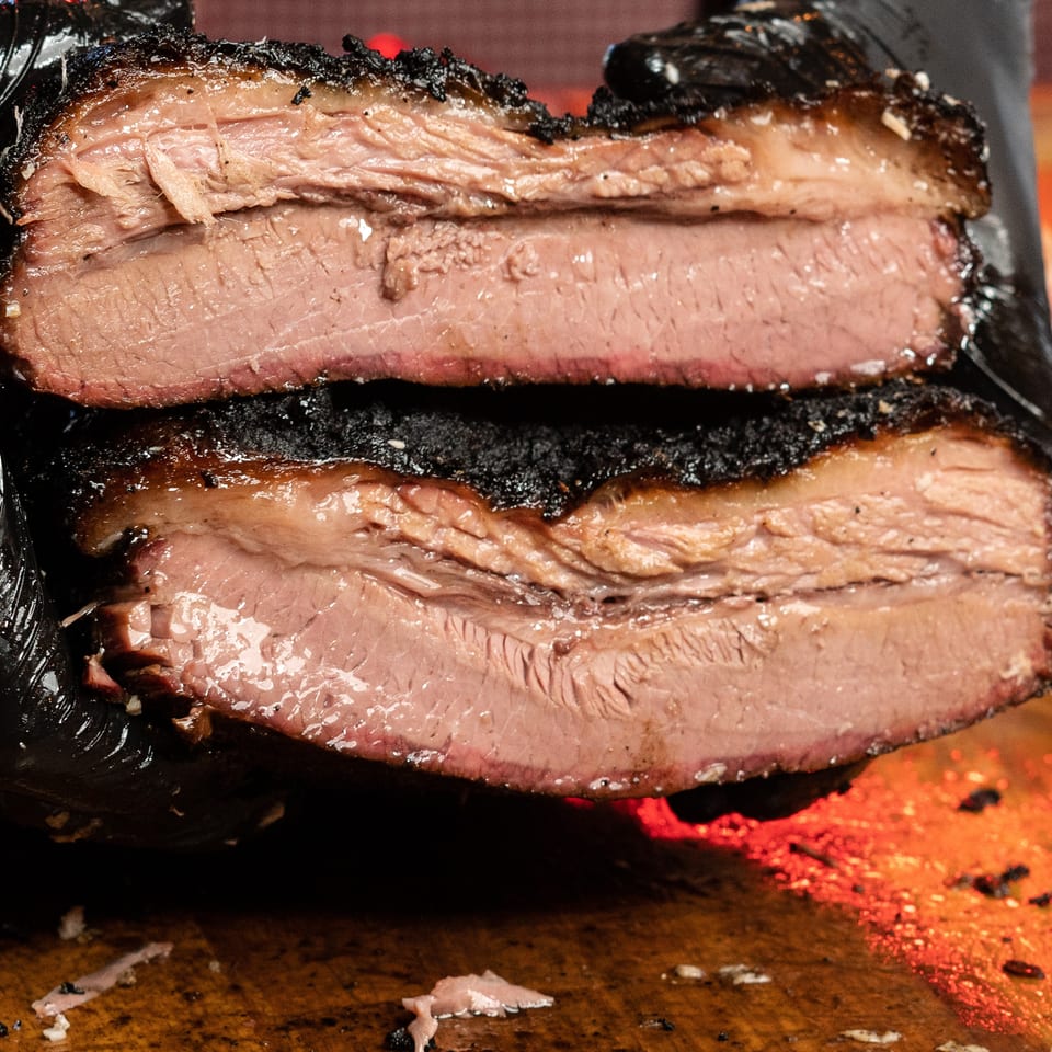 Gloved hand holds two thick slices of smoked brisket, showing juicy pink interiors and dark, crisp bark.