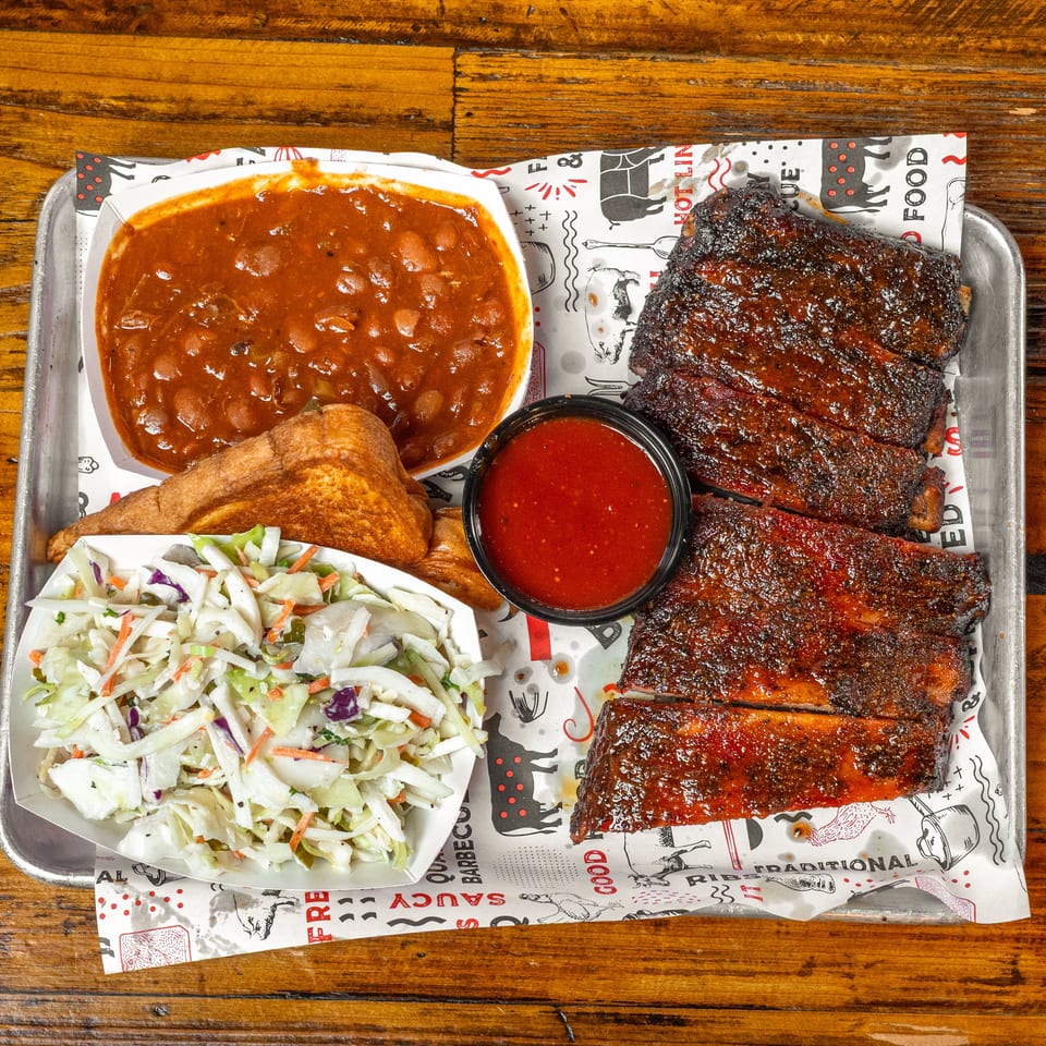 Tray of barbecue ribs with baked beans, coleslaw, cornbread, and red dipping sauce on a wooden table.